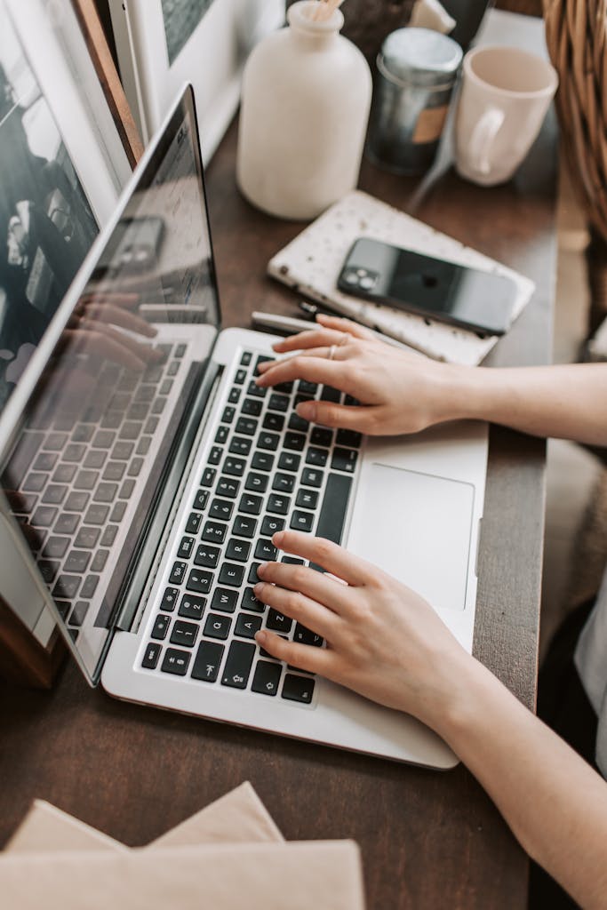 From above of unrecognizable woman sitting at table and typing on keyboard of computer during remote work in modern workspace